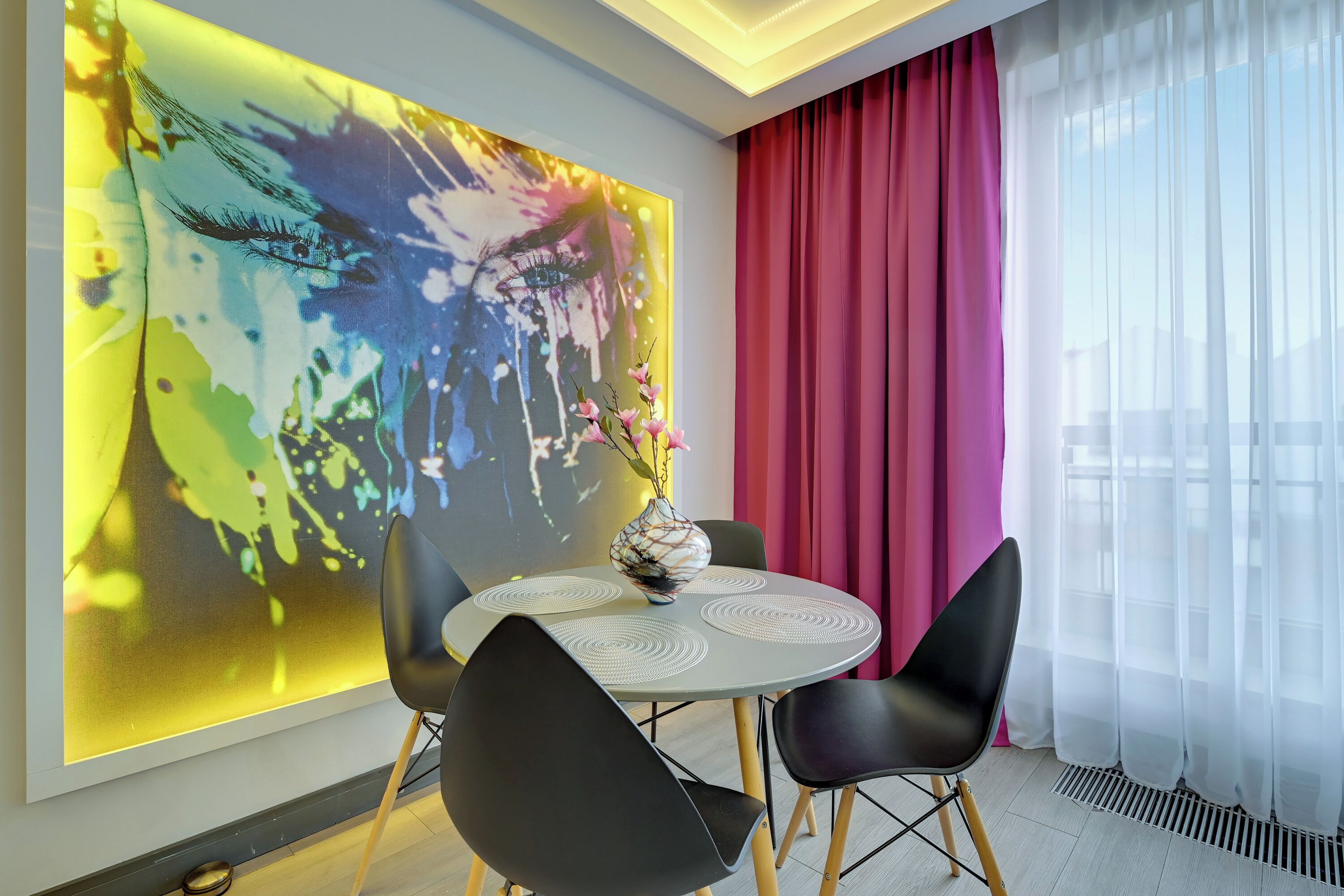 A modern dining area with a round table, two black chairs, a vase with flowers, and a colorful abstract painting on the wall.