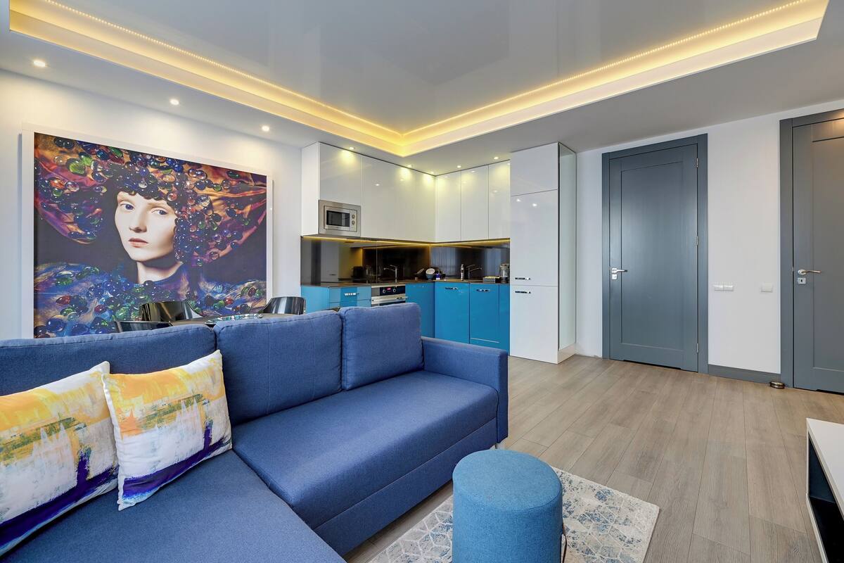 A modern living room with a blue sofa, a painting on the wall, and a kitchen area with blue cabinets.