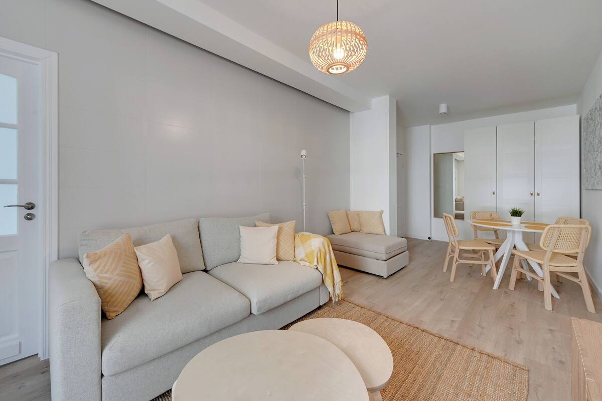 A modern living room with a grey sofa, round coffee tables, and a dining area with wooden chairs and a white table.