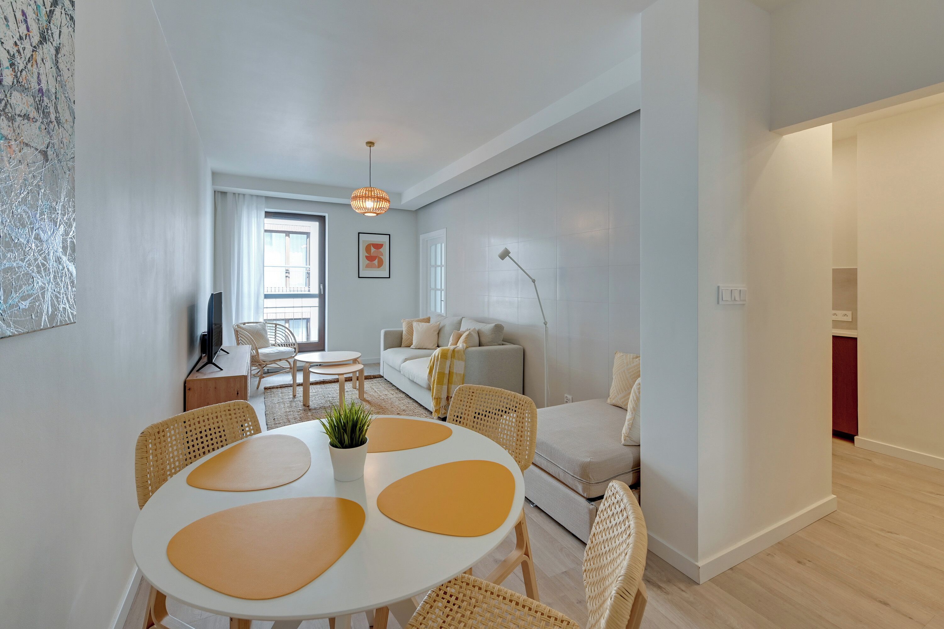 Uma sala de estar moderna com um sofá branco, uma mesa de jantar com jogos americanos amarelos, cadeiras de vime e uma janela grande.