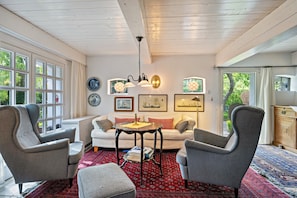 A room with a white sofa, two armchairs, a small table, and a red patterned rug. There are windows, a ceiling fan, and wall-mounted light fixtures. - Vacation home 'Poppenstuuv', idyllic baroque fairytale garden, sauna, parking, Wi-Fi (Stadum)