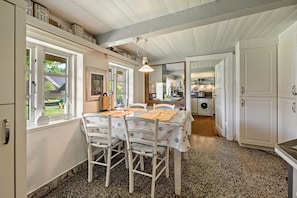 A dining area with a table set for four, chairs, a hanging light fixture, and a view of the kitchen and laundry area. - Vacation home 'Poppenstuuv', idyllic baroque fairytale garden, sauna, parking, Wi-Fi (Stadum)