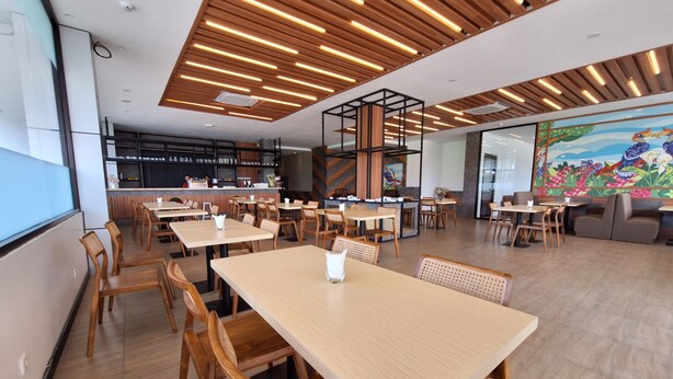 A modern restaurant with wooden tables and chairs, a high ceiling with linear lighting, and a colorful mural on the wall. - Alltrue Hotel Palangkaraya (Palangkaraya)