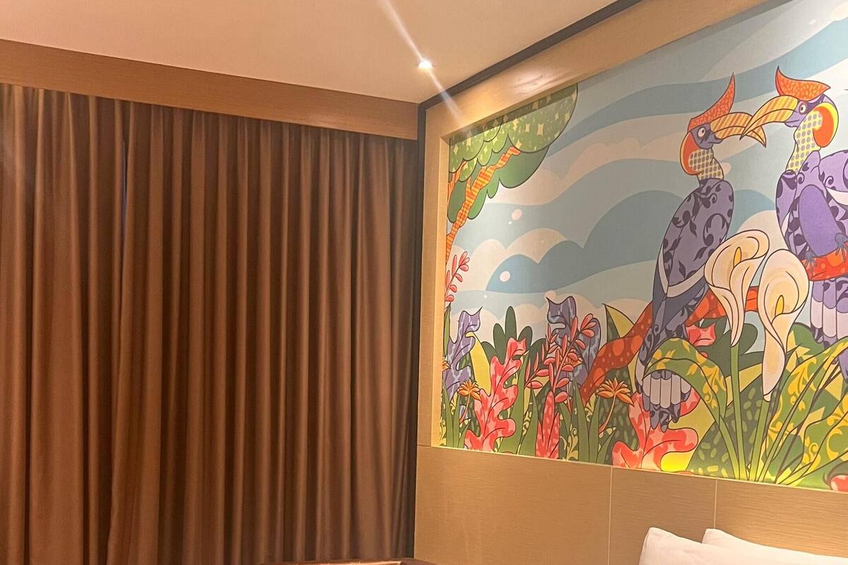 Une chambre d’hôtel avec un grand lit, une chaise, une fresque représentant des oiseaux et des fleurs, et une fenêtre avec des rideaux.