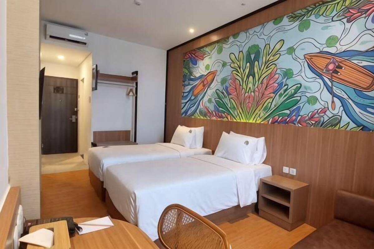 Une chambre d’hôtel avec deux lits, un bureau avec une chaise, une petite table et une fresque colorée sur le mur.