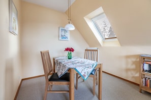 A dining area with a table, chairs, a vase with flowers, a ceiling light, a bookshelf, and a window with blinds.
