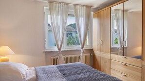 A bedroom with a bed, a bedside lamp, a window with curtains, a radiator, and a wardrobe.