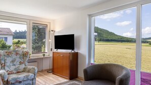 A room with a TV on a wooden cabinet, a floral armchair, a grey armchair, and large windows with a view of a green landscape.