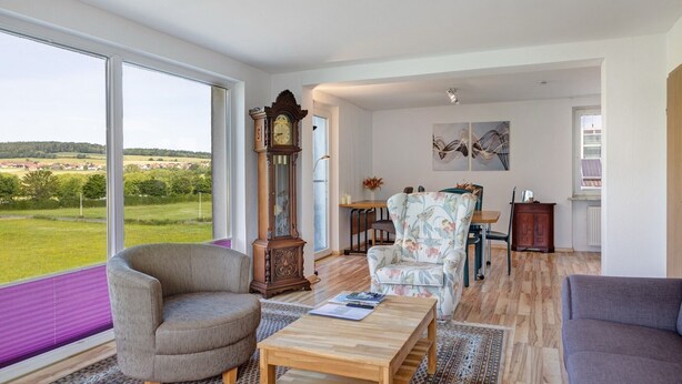 A spacious living room with a large window, a wooden coffee table, a floral armchair, and a classic grandfather clock.