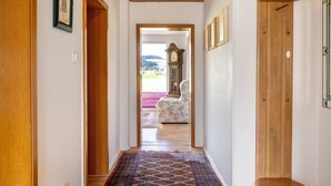 A hallway with a wooden floor, a patterned rug, a wooden door, and a view of a room with a chair and a clock.
