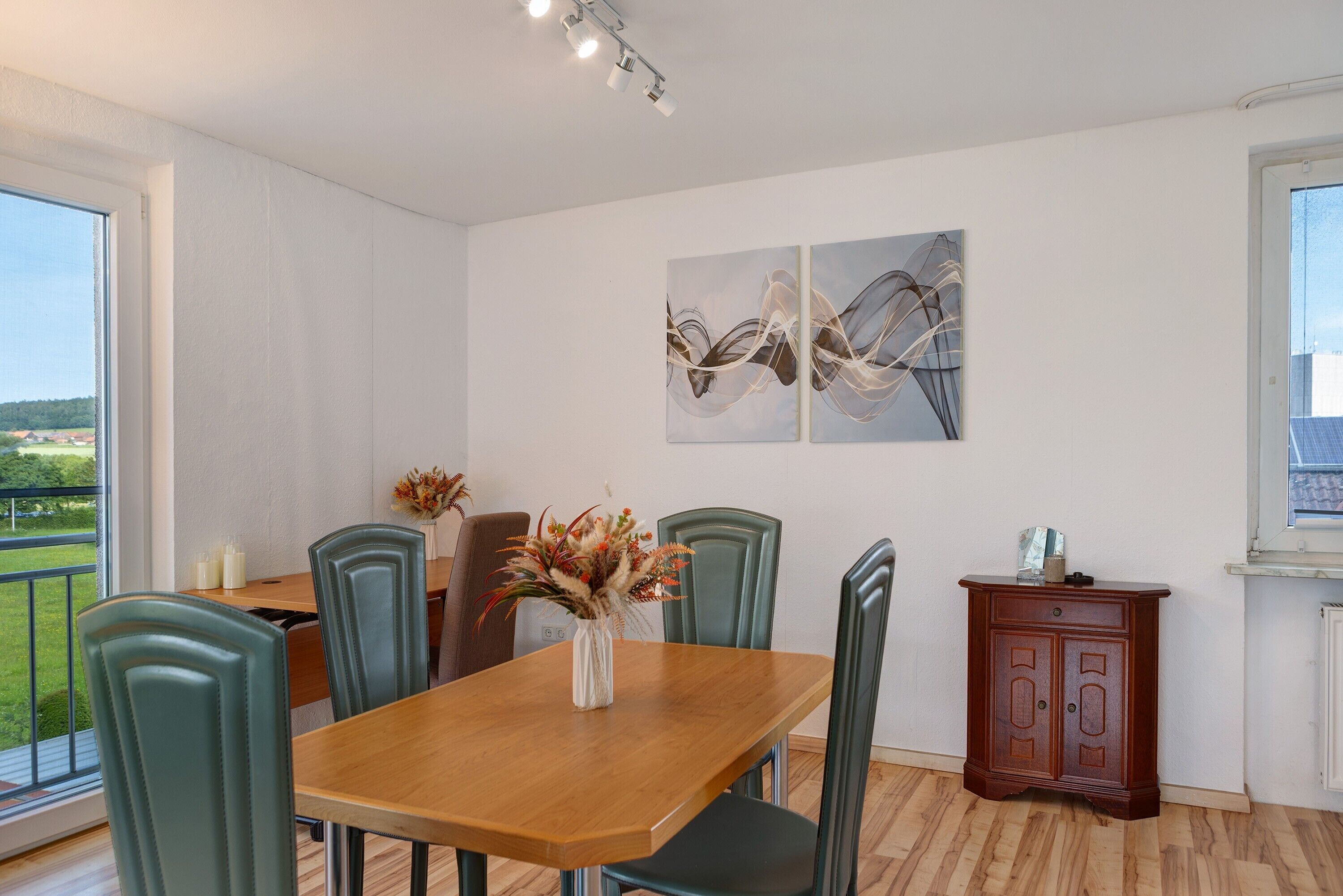 Een eethoek met een houten tafel, groene stoelen, een vaas met bloemen en een kast.