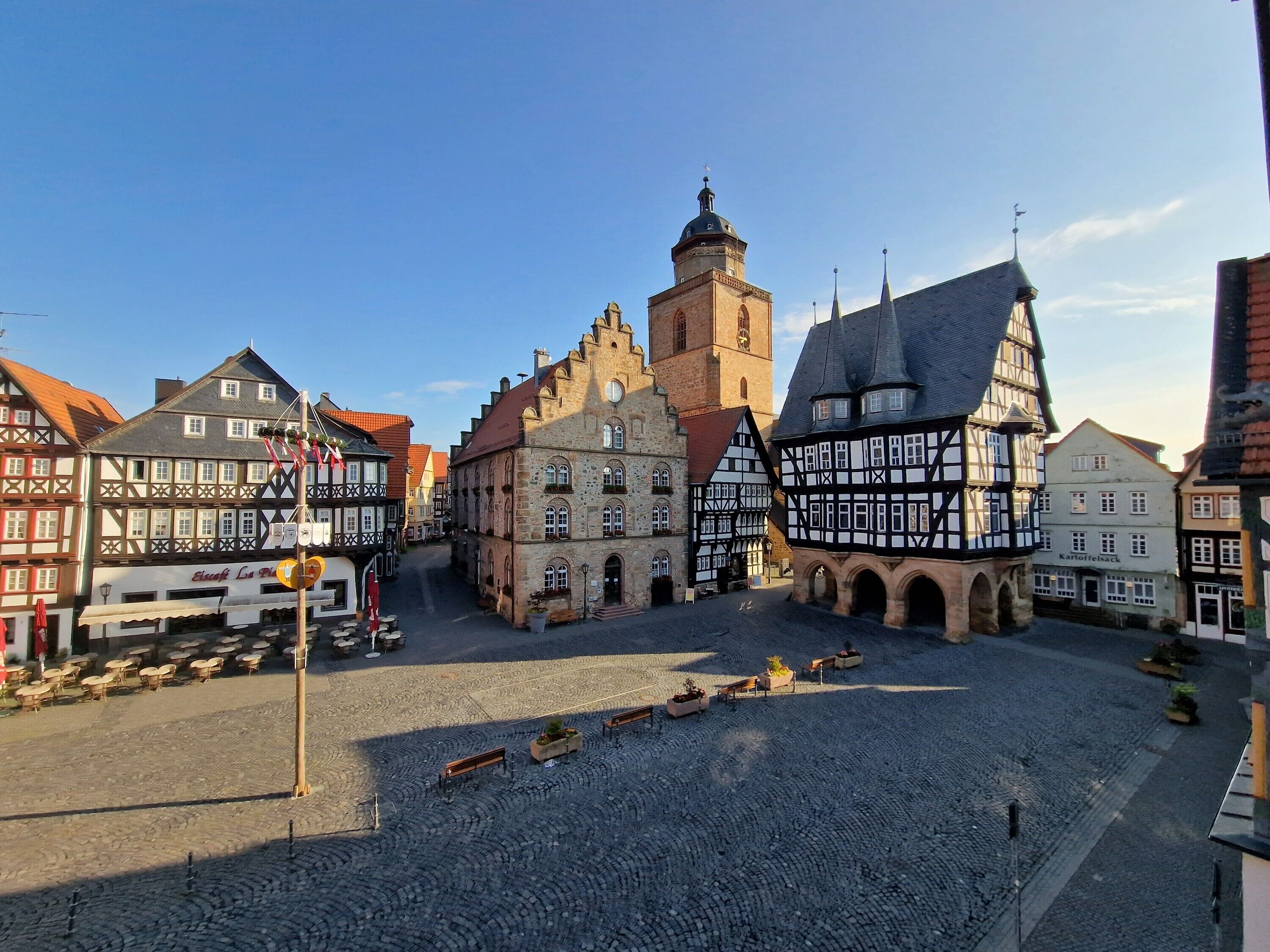 A cobblestone square with traditional half-timbered buildings, a church tower, and a clear sky.