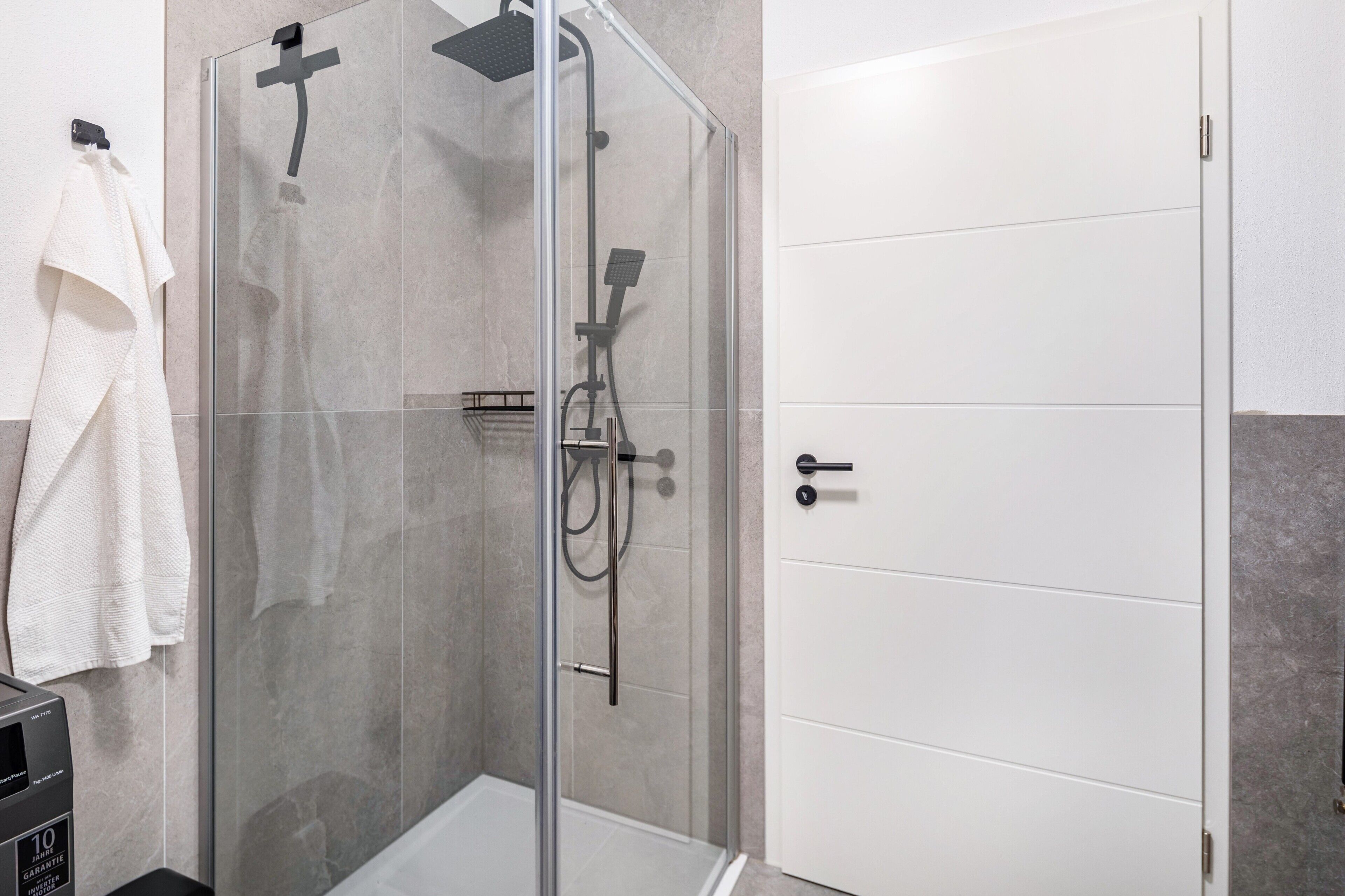 Une douche avec une porte en verre, une serviette blanche accrochée à un crochet et une porte blanche avec une poignée.