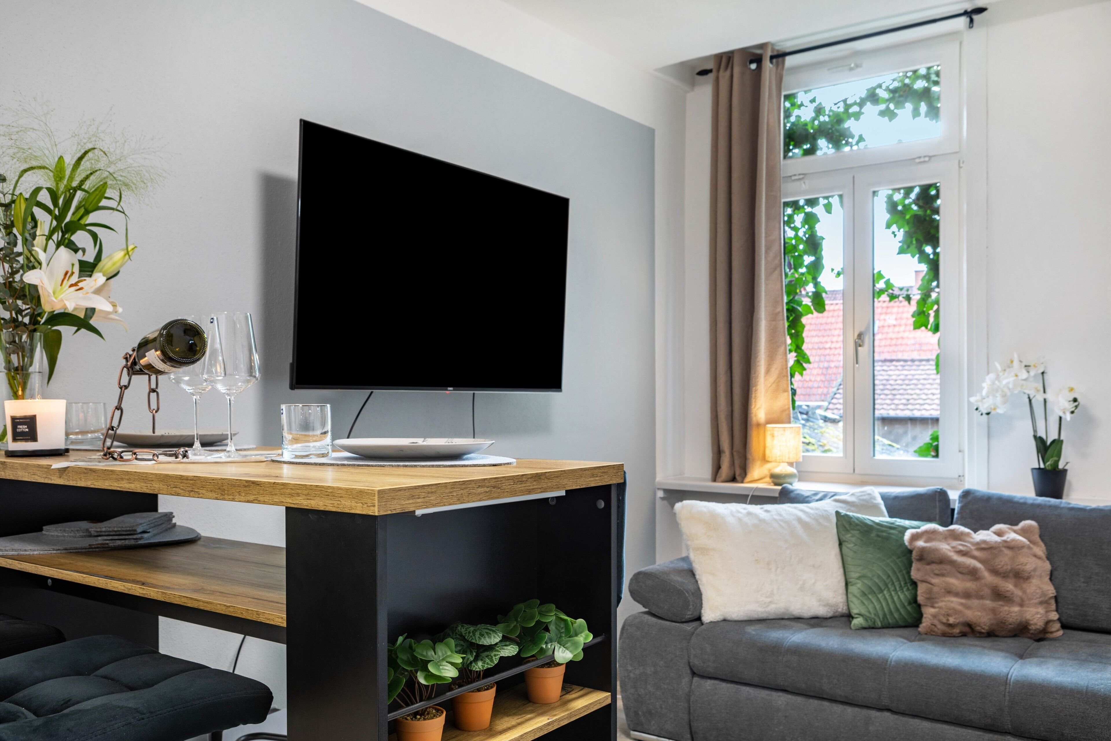 A modern living room with a flat-screen TV, a wooden coffee table, a grey sofa, and a window with curtains.