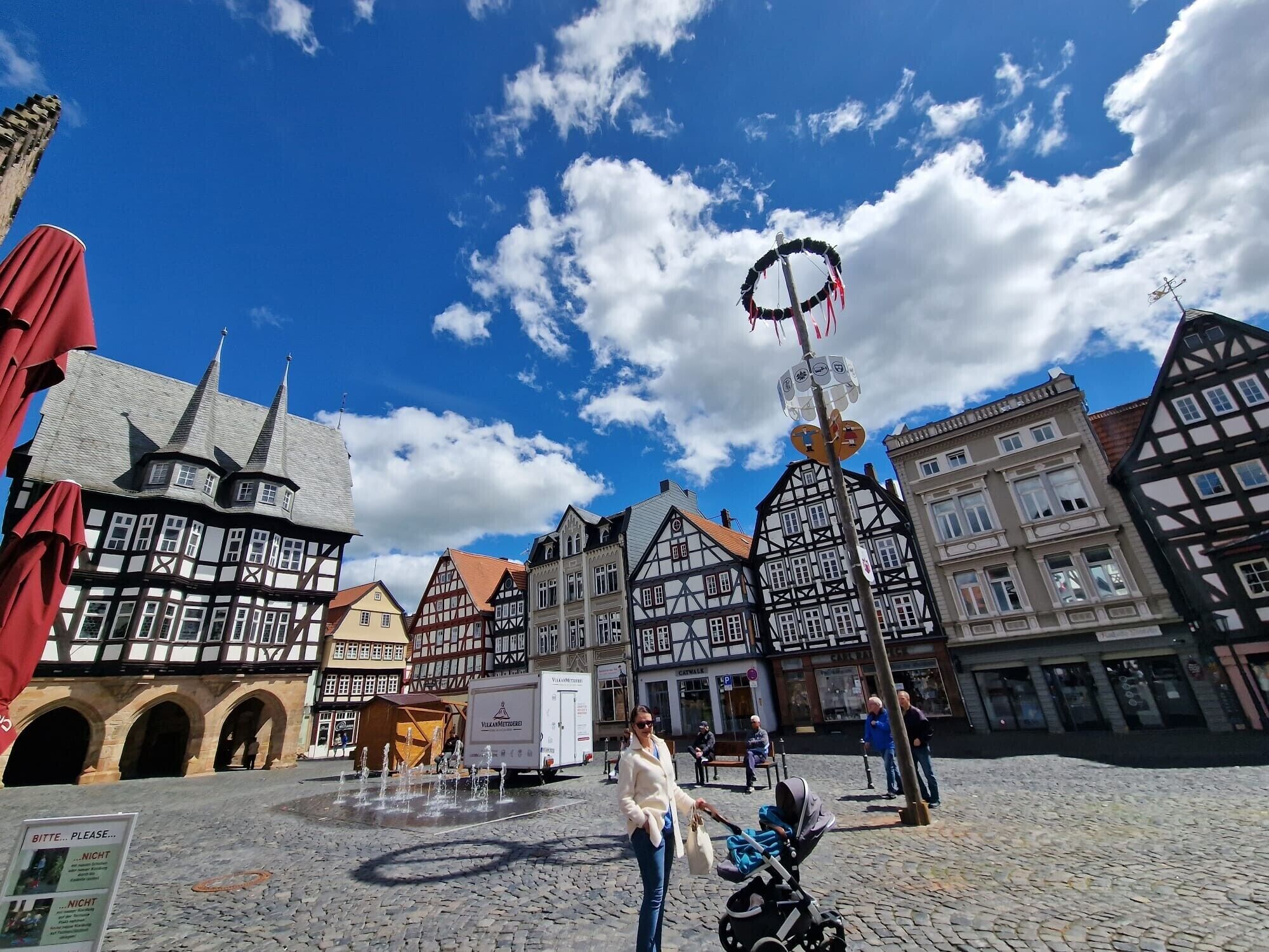A cobblestone square with historic buildings, a fountain, and a signboard.