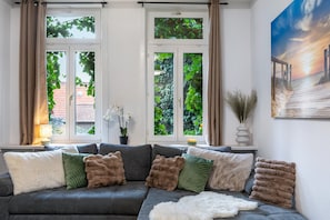 Un salon avec un canapé gris, de nombreux coussins, un tapis blanc à longues mèches et un tableau représentant un coucher de soleil sur une jetée.