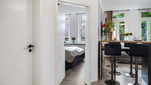 A modern interior with a kitchen area, a dining table with bar stools, and a visible bedroom with a bed and window. - Apartment 'Marktplatz Ferienwohnung' with Wi-Fi (Alsfeld)