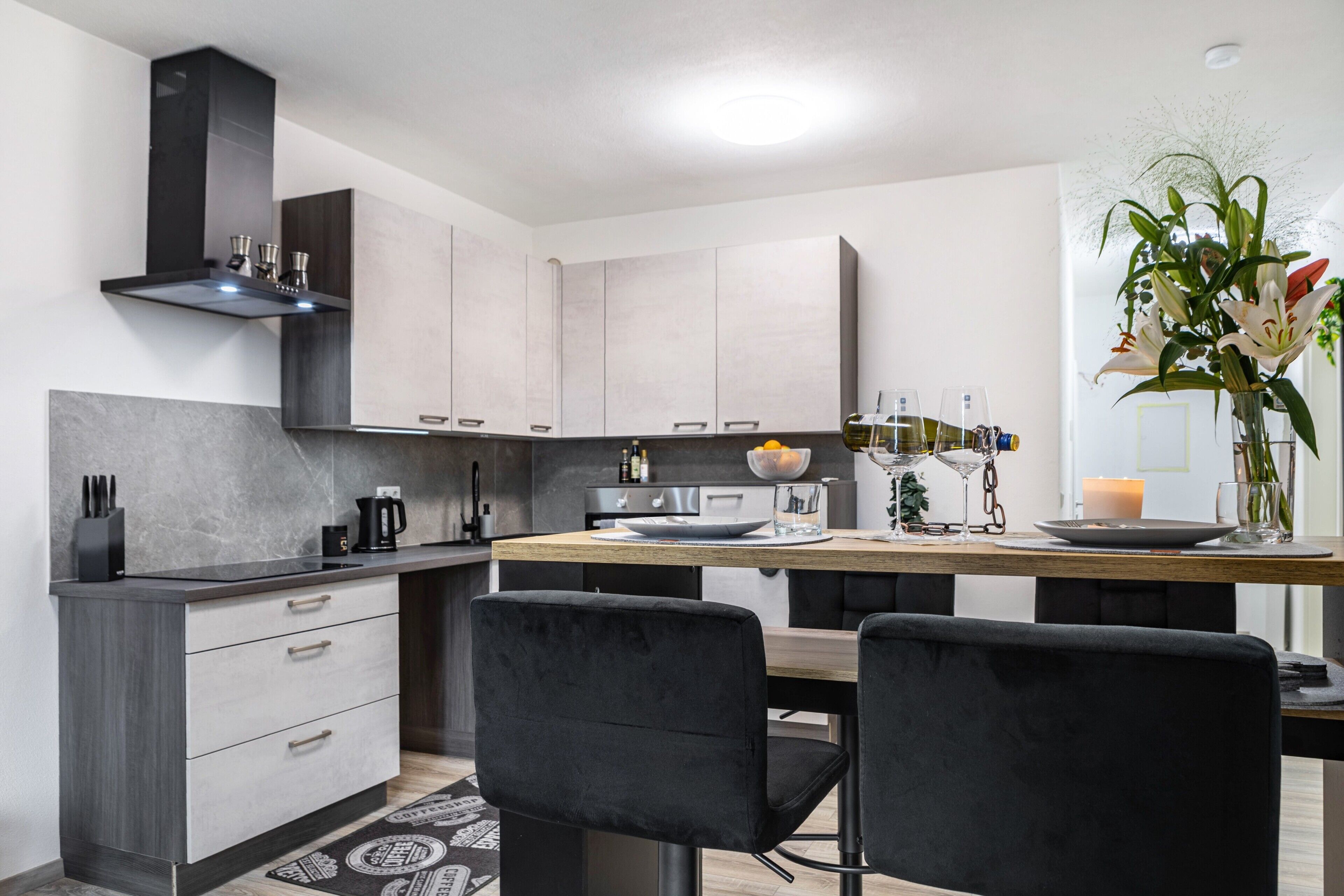 A modern kitchen with grey cabinets, a black range hood, and a dining area with two black chairs.