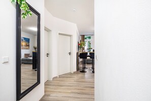 Un couloir avec un grand miroir, un plancher en bois, un coin repas avec des chaises et une table, et une porte donnant sur une autre pièce.