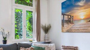 A living room with a sofa, a window with curtains, a painting of a beach scene, and a potted plant. - Apartment 'Marktplatz Ferienwohnung' with Wi-Fi (Alsfeld)