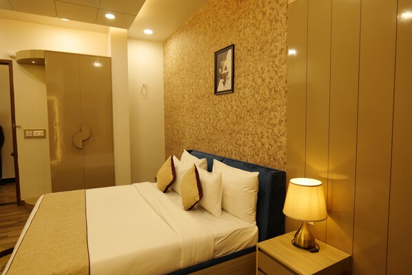 A hotel room with a bed, a nightstand, a lamp, a framed picture on the wall, and a door. - Tathastu Wilderness by The Kosi River (Ramnagar)
