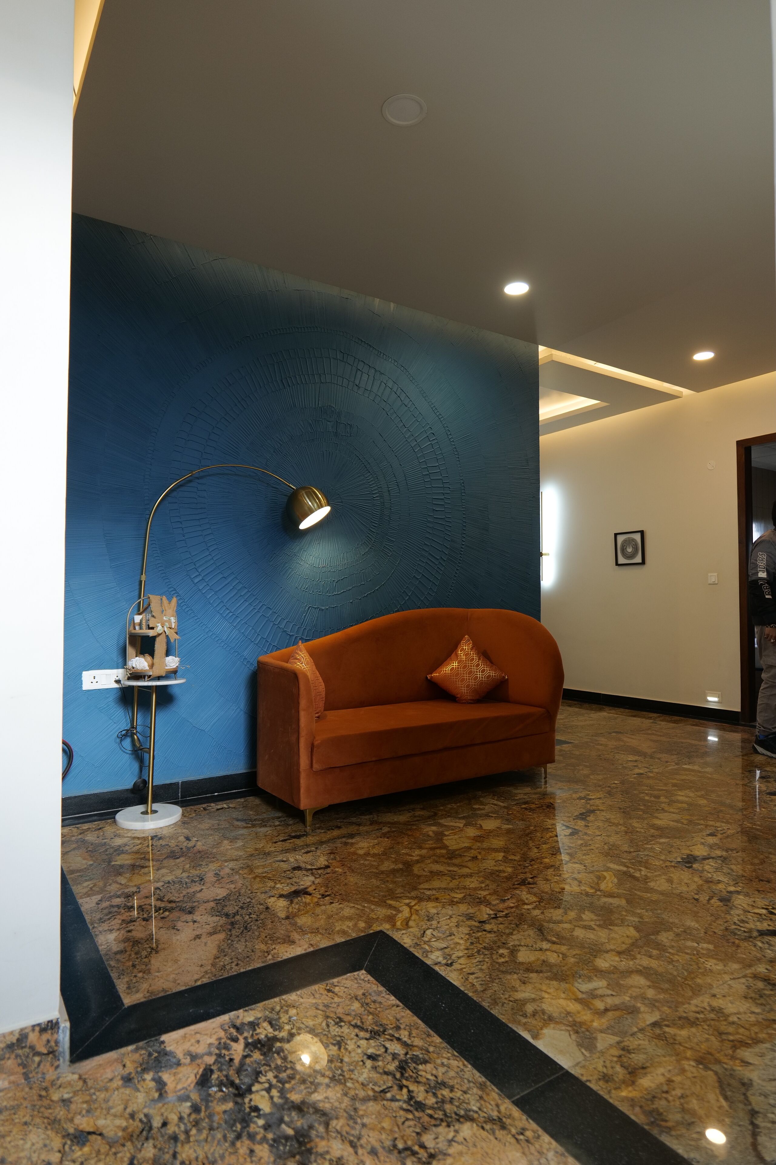 Eine moderne Lobby mit einer blauen Wand mit Struktur, einem braunen Sofa mit gemustertem Kissen, einer Stehlampe und einem Marmorboden.