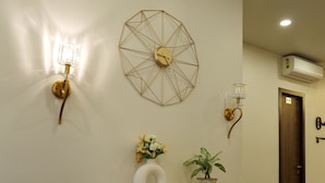 Un pasillo con suelo de mármol, un aplique geométrico en la pared, una planta en maceta y un jarrón blanco con flores.