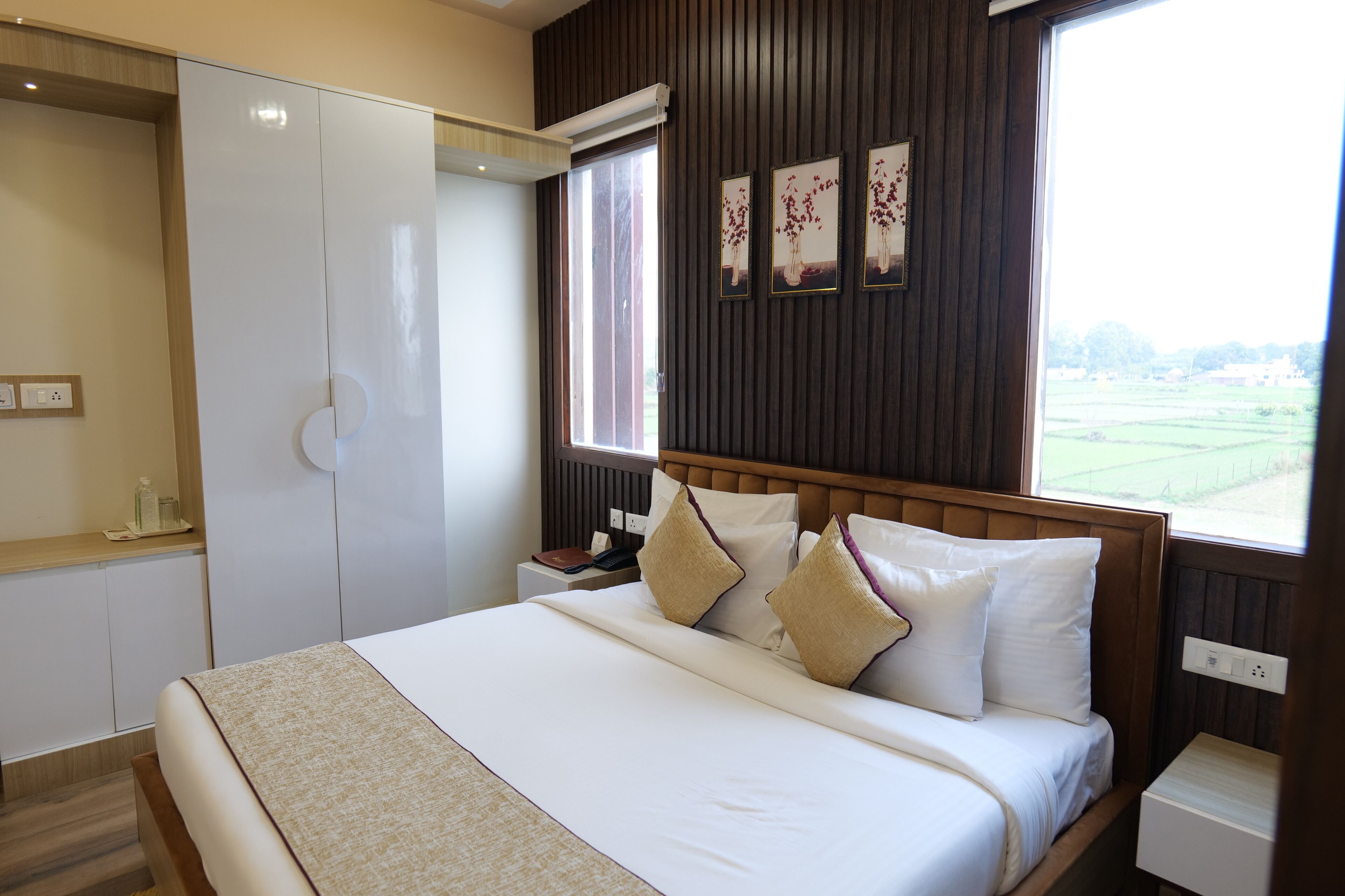 A neatly made bed with a white comforter and two decorative pillows, a wooden headboard, a bedside table with a lamp, and a window with a view of a green landscape.