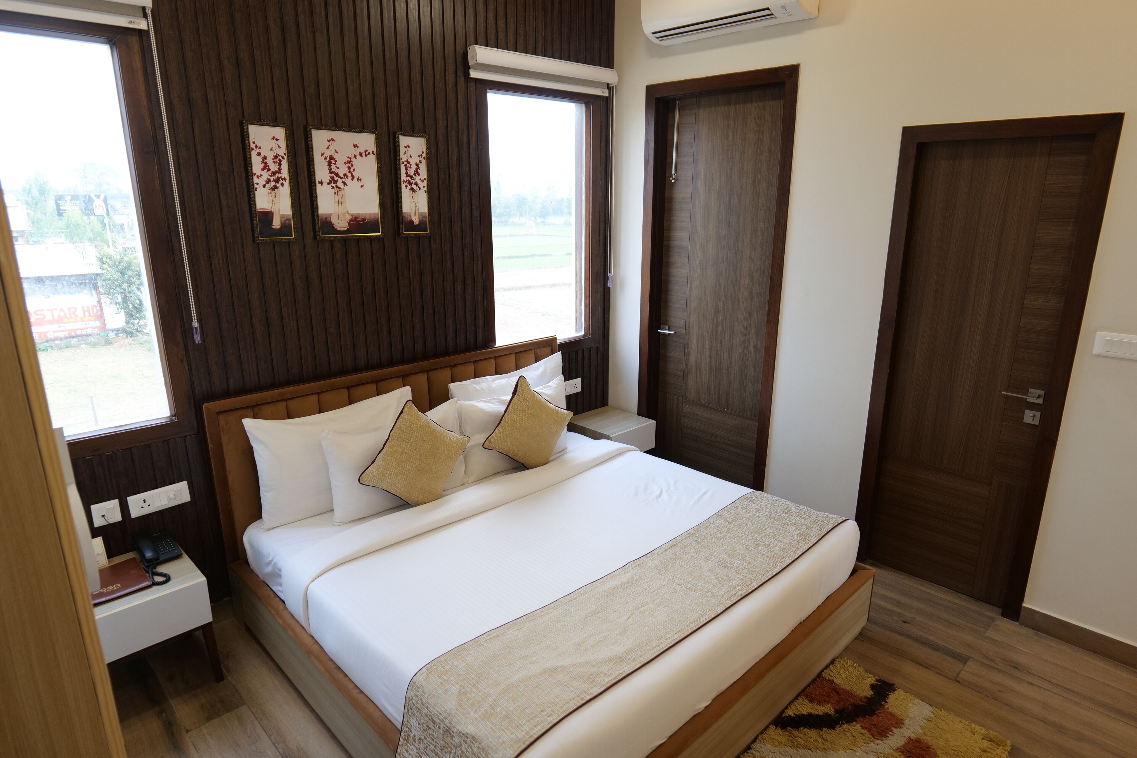 A neatly arranged bedroom with a wooden headboard, a large bed with white linens, a bedside table with a phone and a book, and a window with a view of greenery.