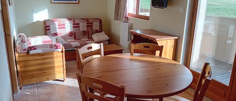 Una stanza accogliente con un tavolo e sedie in legno, un divano con cuscini decorati, una televisione a parete e una finestra che si affaccia su un paesaggio verde.