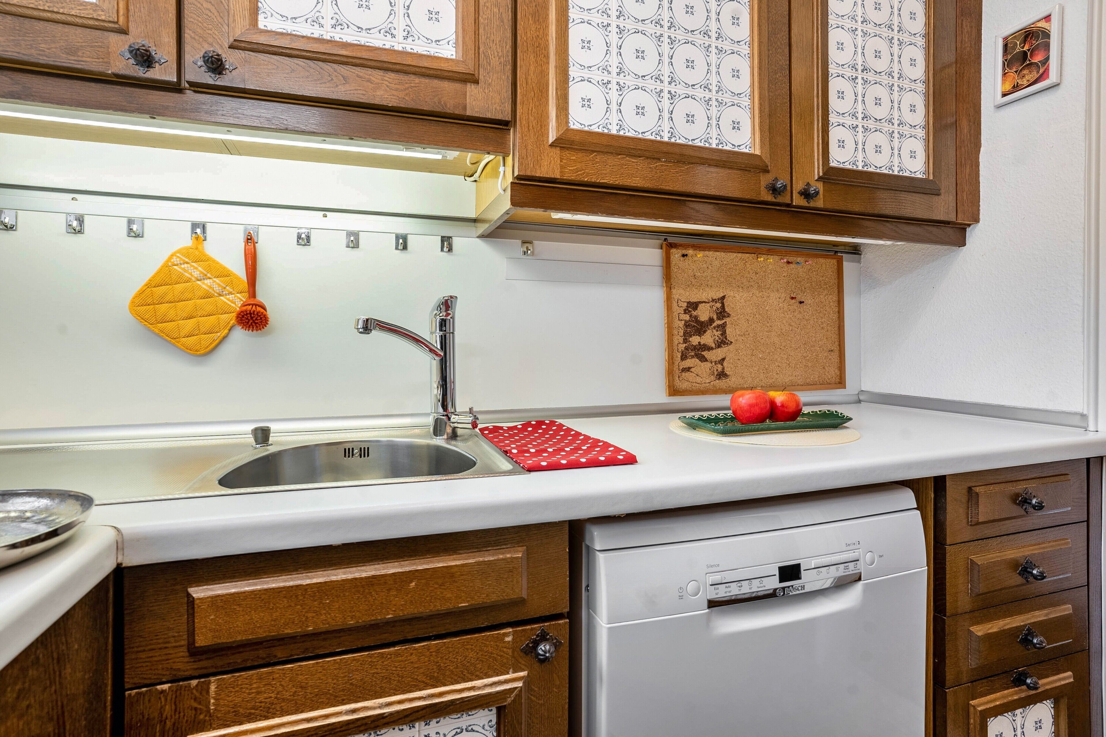 A kitchen with wooden cabinets, a stainless steel sink, a dishwasher, and a corkboard with notes.