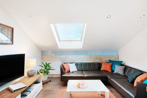 A modern living room with a large flat-screen TV, a leather sofa, a white coffee table, and a skylight.