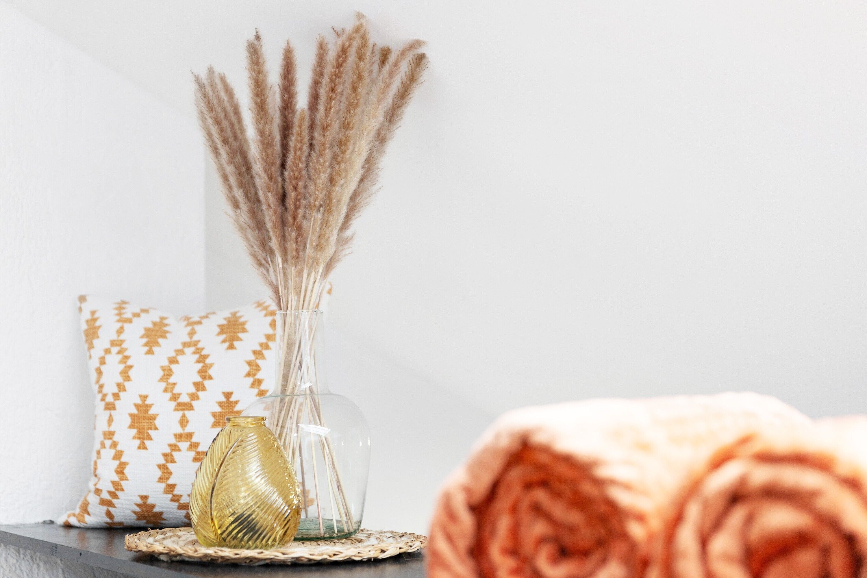 A clear glass vase with dried pampas grass, a patterned pillow, and a rolled-up beige blanket on a woven mat.