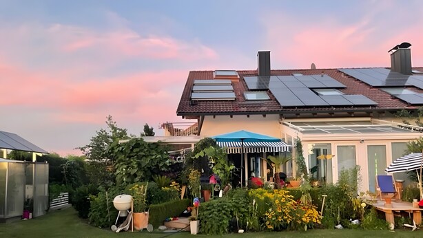 Una casa moderna con paneles solares, un jardín con diversas plantas, un patio con una sombrilla a rayas y un invernadero.