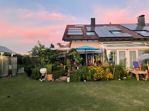 A modern house with solar panels, a garden with various plants, a patio with a striped umbrella, and a conservatory.