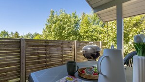 Une terrasse avec une clôture en bois, un barbecue, une table avec un magazine, une carafe et un panier de fruits.