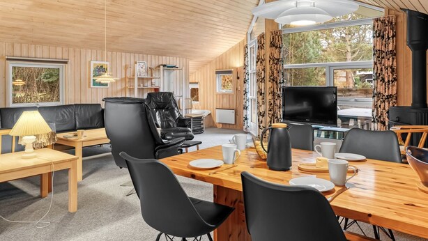 L’intérieur d’un chalet accueillant, avec un plafond en bois, une table à manger dressée pour quatre personnes, un canapé en cuir noir et une grande fenêtre agrémentée de rideaux.