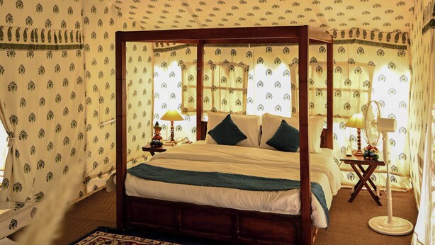 A four-poster bed with a floral-patterned canopy, a patterned rug, and a small table with a lamp. - Bookmark Shivir (Phulpur)