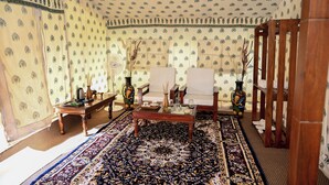 A room with patterned wallpaper, a large patterned rug, two chairs, a wooden coffee table, and a decorative vase. - Bookmark Shivir (Phulpur)