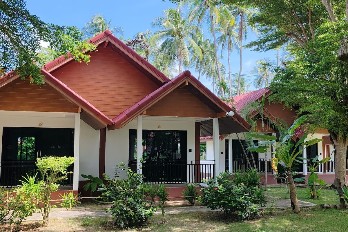 A bungalow-style building with a red roof and white walls, surrounded by lush greenery and palm trees.