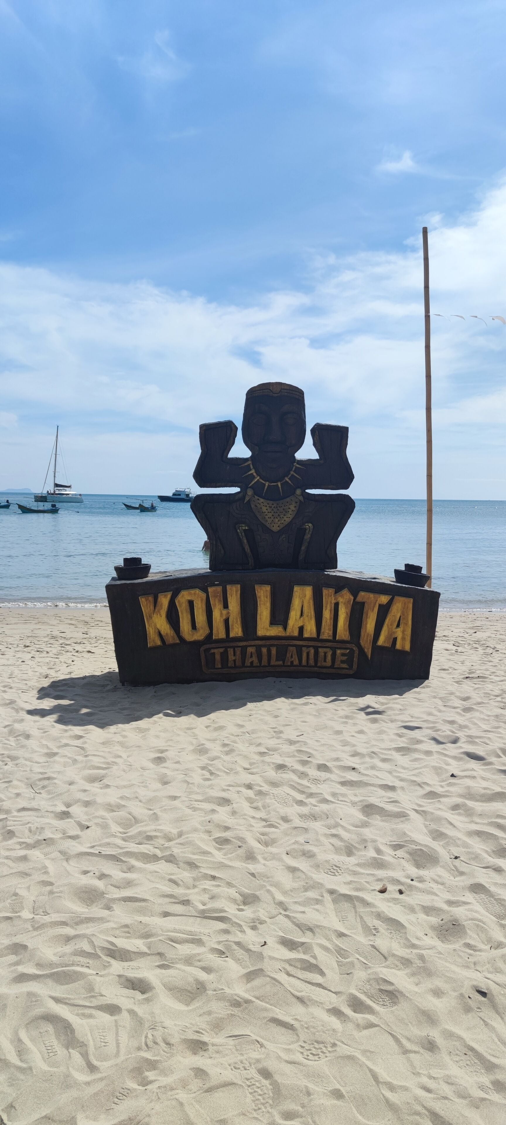 A wooden sign with the text "Koh Lanta Thailand" on a sandy beach with boats in the background.