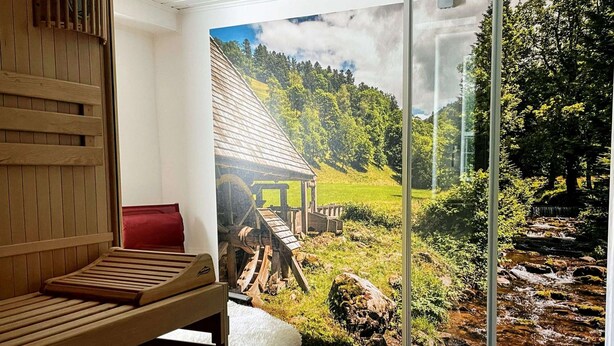 A sauna room with a wooden bench, a red cushion, and a glass door leading to a scenic outdoor view of a wooden structure, a stream, and a forested hillside. - Vacation home Hirschle -Sauna, dogs, 3 bedrooms, 8 persons (Alpirsbach)