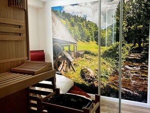 A sauna room with a wooden bench, a red cushion, and a glass door leading to a scenic outdoor view of a wooden structure, a stream, and a forested hillside. - Vacation home Hirschle -Sauna, dogs, 3 bedrooms, 8 persons (Alpirsbach)