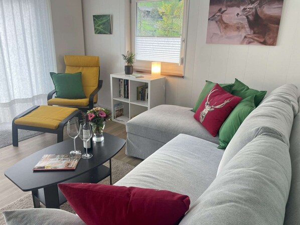 A living room with a sofa, armchair, and coffee table. There is a bookshelf and a window with blinds. - Vacation home Hirschle -Sauna, dogs, 3 bedrooms, 8 persons (Alpirsbach)