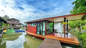 A modern floating house with a wooden deck, glass windows, and a covered outdoor area. - Boathouse Boutique Riverside (Nakhon Chai Si)