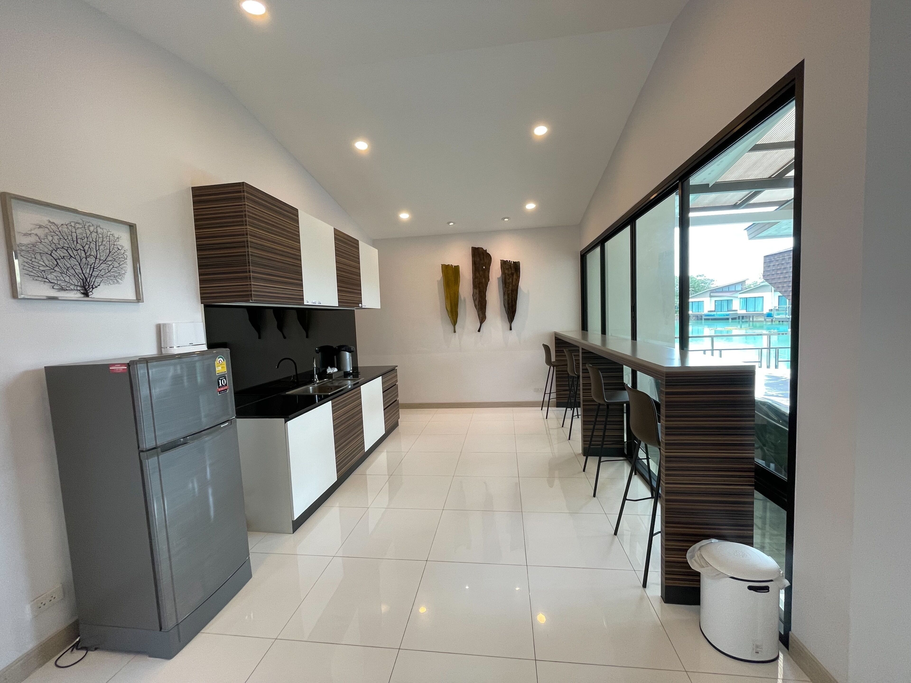 Una cocina moderna con refrigerador, estufa y lavaplatos. Hay un comedor con sillas y una mesa. La habitación tiene ventanas amplias y vista a una piscina.