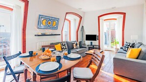 A modern living room with a dining area, featuring a sofa, a TV, and a breakfast setup with plates, cups, and a fruit bowl. - Bica's River View Duplex (Lisbon)