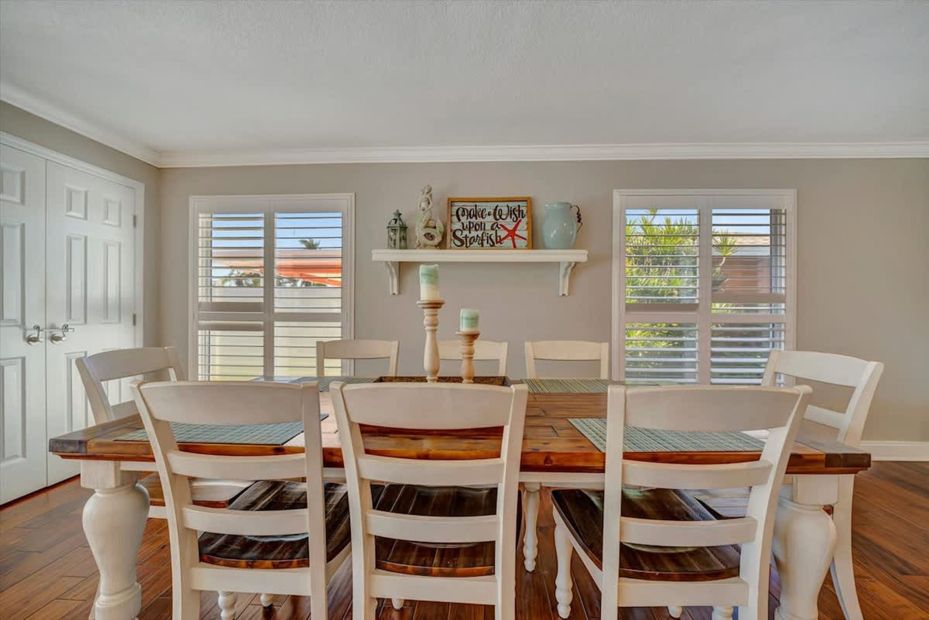 A dining room with a wooden table and white chairs, a decorative sign on the wall, and a window with blinds.