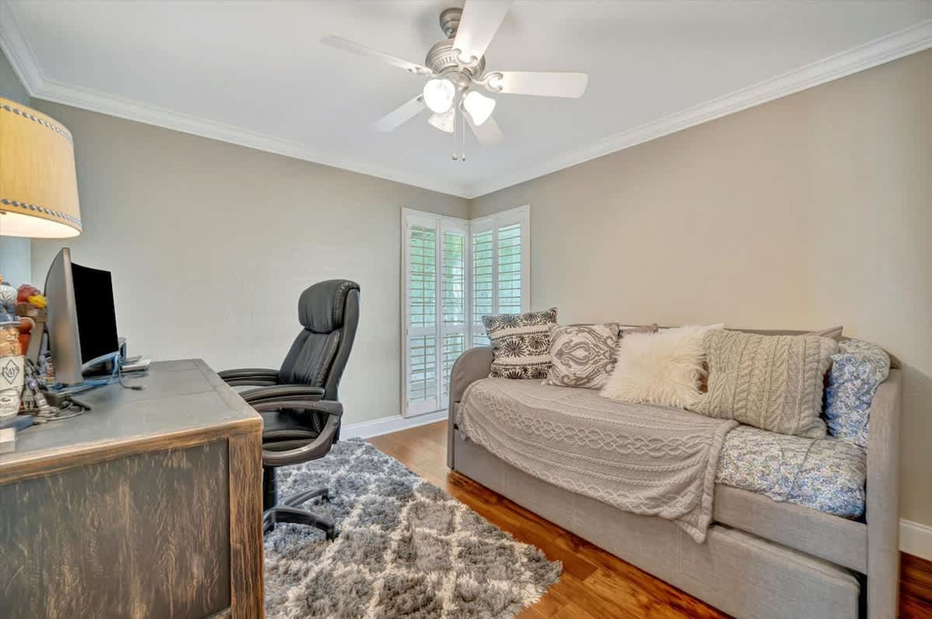 A bedroom with a bed, a desk, a chair, a ceiling fan, and a window with shutters.