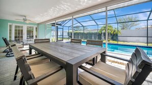 Un patio iluminado por el sol con piscina, una mesa de madera y sillas con cojines.
